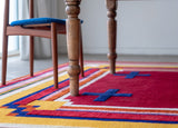 close up of a mostly red rug with blue and yellow border with two blue crosses under a wood table with one chair