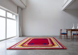 Mostly red rug with yellow and blue striped border and four blue crosses sitting on the floor next to a wood table with two chairs in a white room 