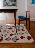 Light pink rug with red and light blue crosses sitting on a wood floor under a wood table with a wood chair