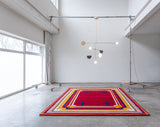 Mostly red rug with yellow and blue border and blue crosses sitting on the floor with a light designed by Anony hanging above in a white room