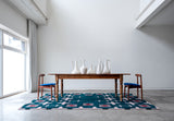 Green rug with a light pink hashtag design sitting on the floor under a wood table with two chairs in a white room