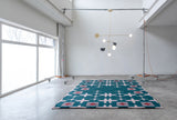 Green rug with a light pink hashtag design sitting on the floor with a light designed by Anony hanging above in a white room