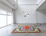 Orange green and yellow brightly designed rug sitting on the floor with a light designed by Anony hanging above in a white room