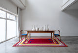 Mostly red rug with blue and yellow border and blue crosses under a wood table with two chairs in a white room