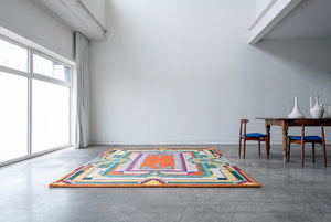 Orange green and yellow brightly designed rug sitting on the floor next to a wood table with two chairs in a white room 