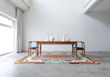 Orange green and yellow brightly designed rug sitting on the floor under a wood table with two chairs in a white room