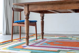 Close up of a orange green and yellow brightly designed rug sitting on the floor under a wood table with one chairs