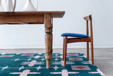 Close up of a green rug with a light pink hashtag design sitting on the floor under a wood table and chair
