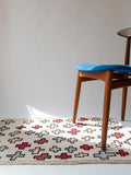 Light pink rug with red and light blue crosses sitting on the floor under a wood chair with a blue seat in a white space